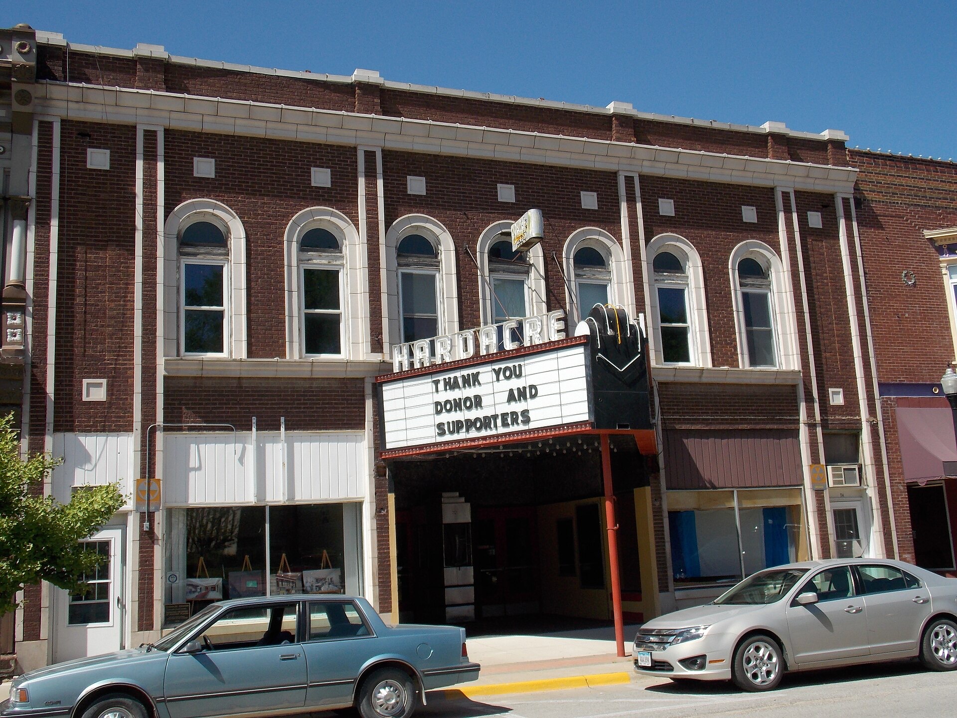 Daytime street view of the Hardacre Theater in Tipton, Iowa, showing its red-brick two-story facade, arched second-story windows, and vintage black and white neon marquee.